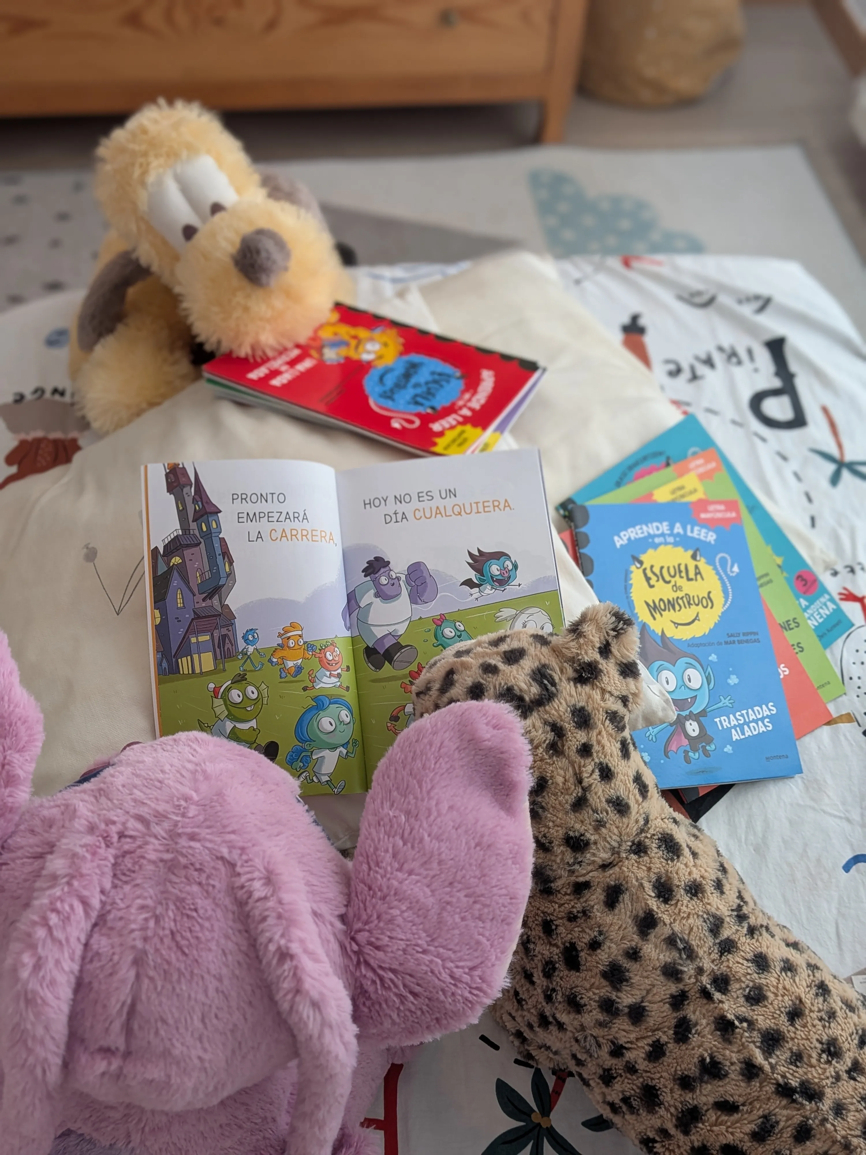 Un libro de la Escuela de Monstruos abierto sobre la cama, rodeado de peluches y del resto de títulos de la colección