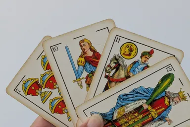Una mano sostiene cuatro cartas de la baraja española en abanico: el siete de copas, la sota de espadas, el caballo de oros y el rey de bastos