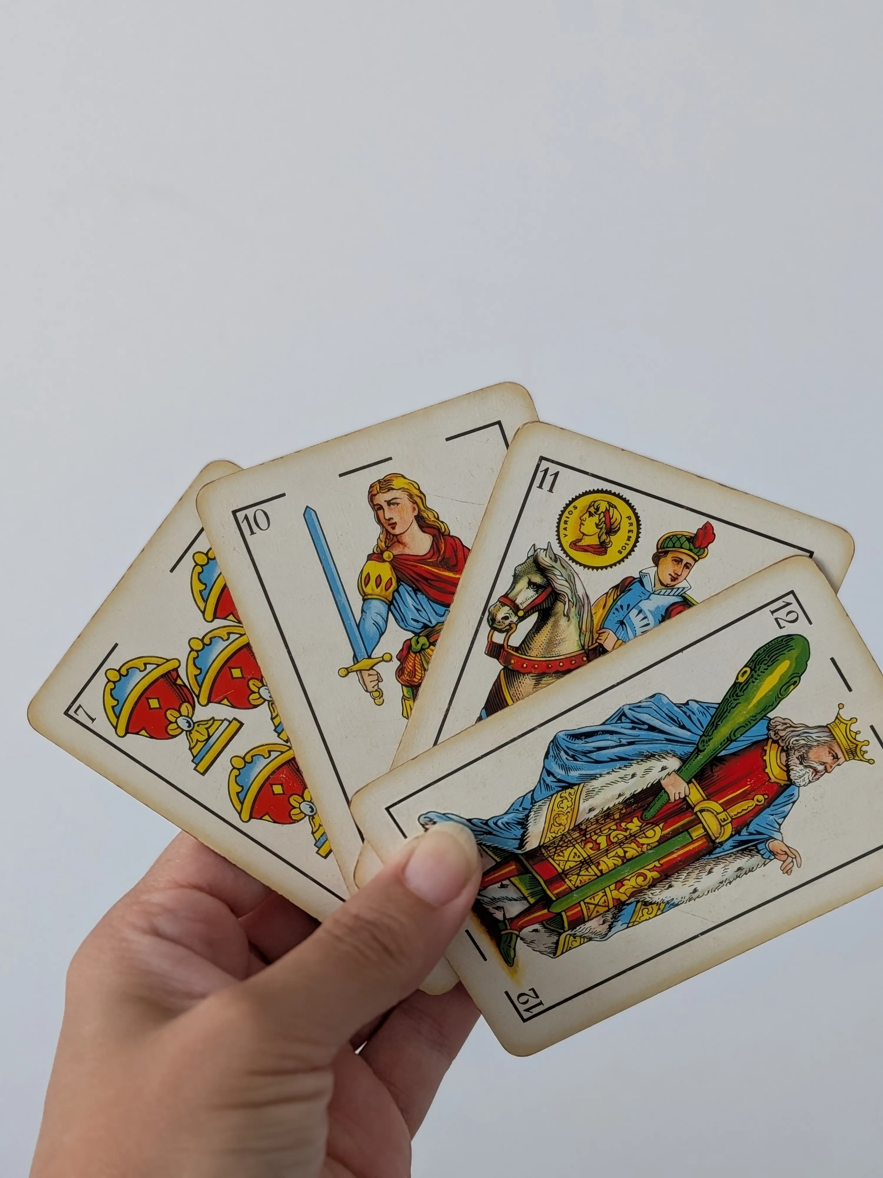 Una mano sostiene cuatro cartas de la baraja española en abanico: el siete de copas, la sota de espadas, el caballo de oros y el rey de bastos