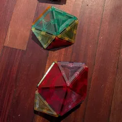 Dos poliedros construidos con piezas magnéticas translúcidas, uno verde y amarillo y otro rojo y rosa, sobre el suelo de madera