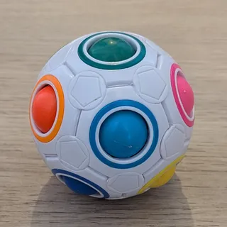 La bola arcoíris de rompecabezas, blanca y del tamaño de una pelota pequeña, con las bolitas verde, naranja, azul, rosa y amarilla asomando por sus huecos