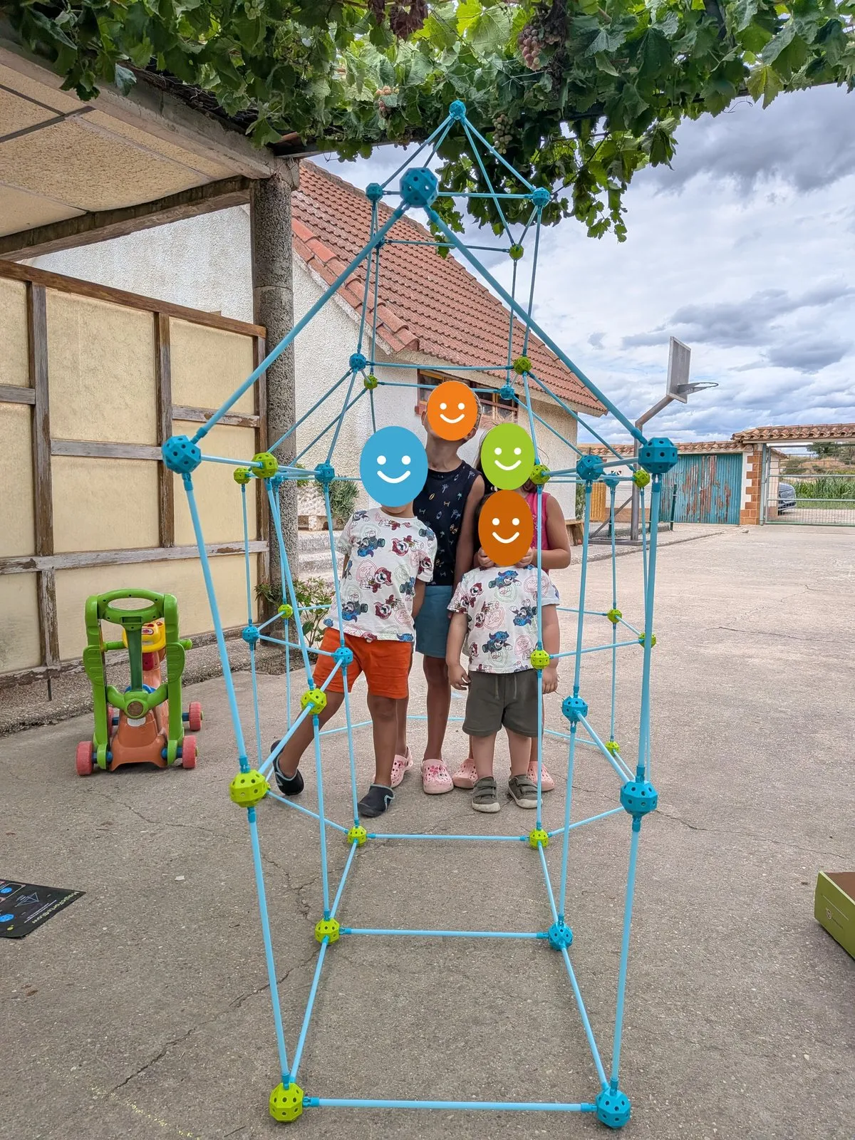 Una cabaña alta montada con varillas azules y conectores de colores en el patio, con cuatro niños de pie dentro