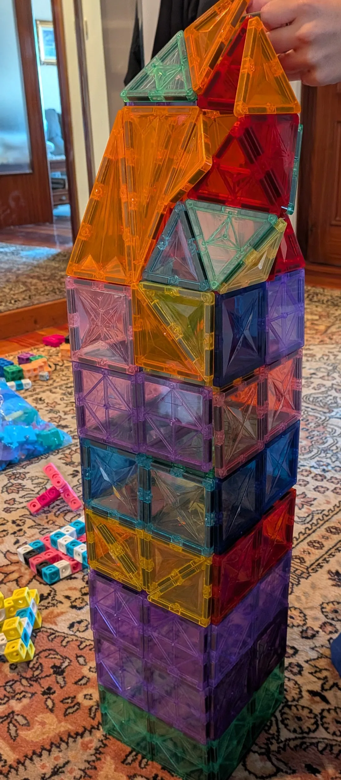Una torre alta construida con piezas magnéticas de colores, con un tejado de triángulos naranjas y rojos, sobre una alfombra