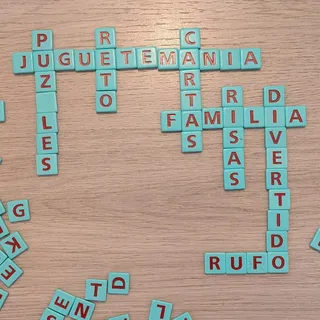 Fichas de letras turquesa formando palabras cruzadas sobre el suelo: juguetemania, puzzles, reto, cartas, familia, risas y divertido