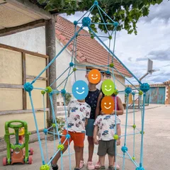 La cabaña montada en el patio con varillas azules y conectores de colores, alta como un adulto, con cuatro niños de pie dentro