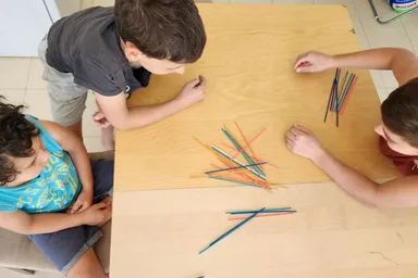 Tres niños jugando al mikado alrededor de una mesa de madera, vistos desde arriba