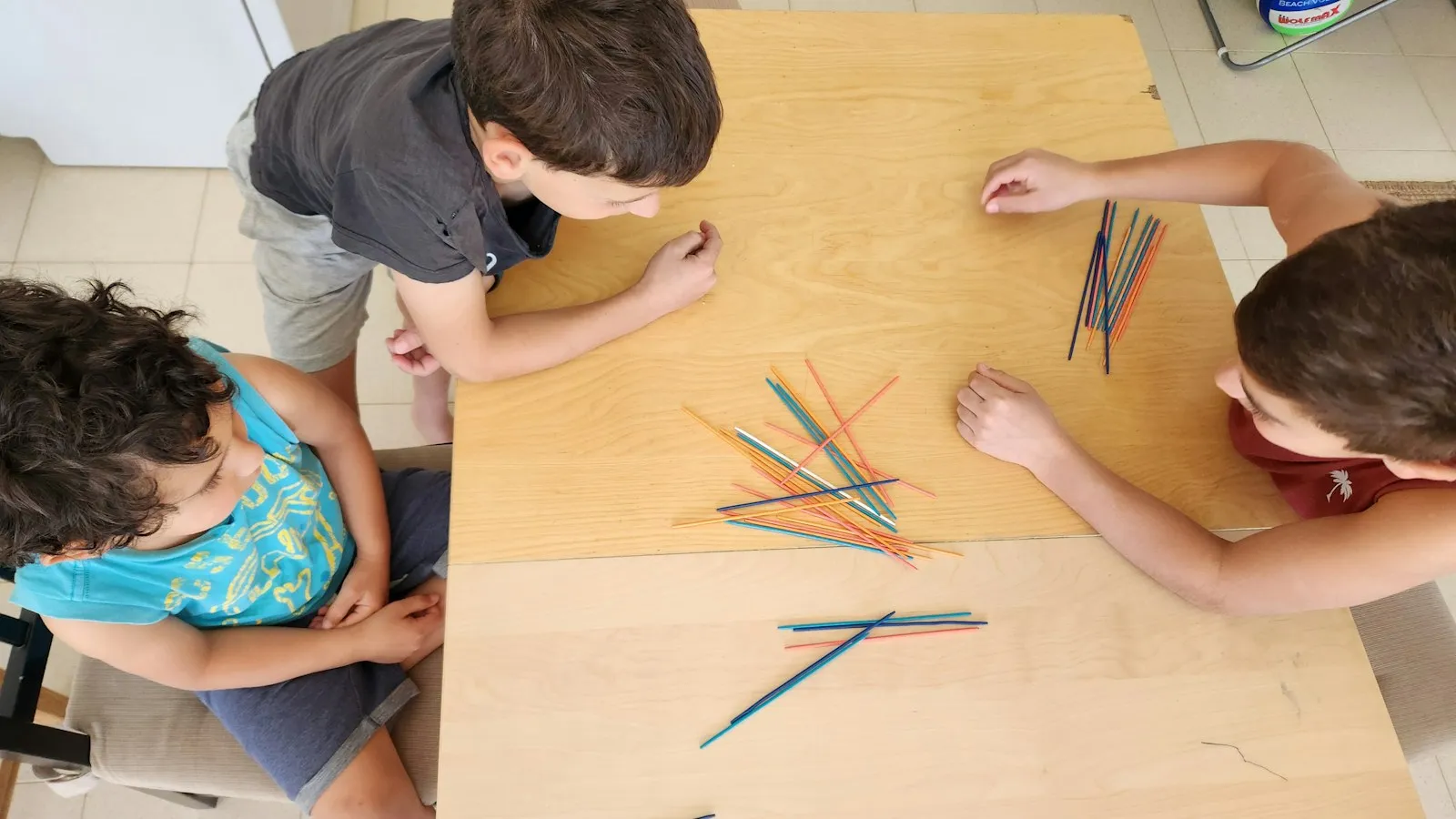 Tres niños jugando al mikado alrededor de una mesa de madera, vistos desde arriba