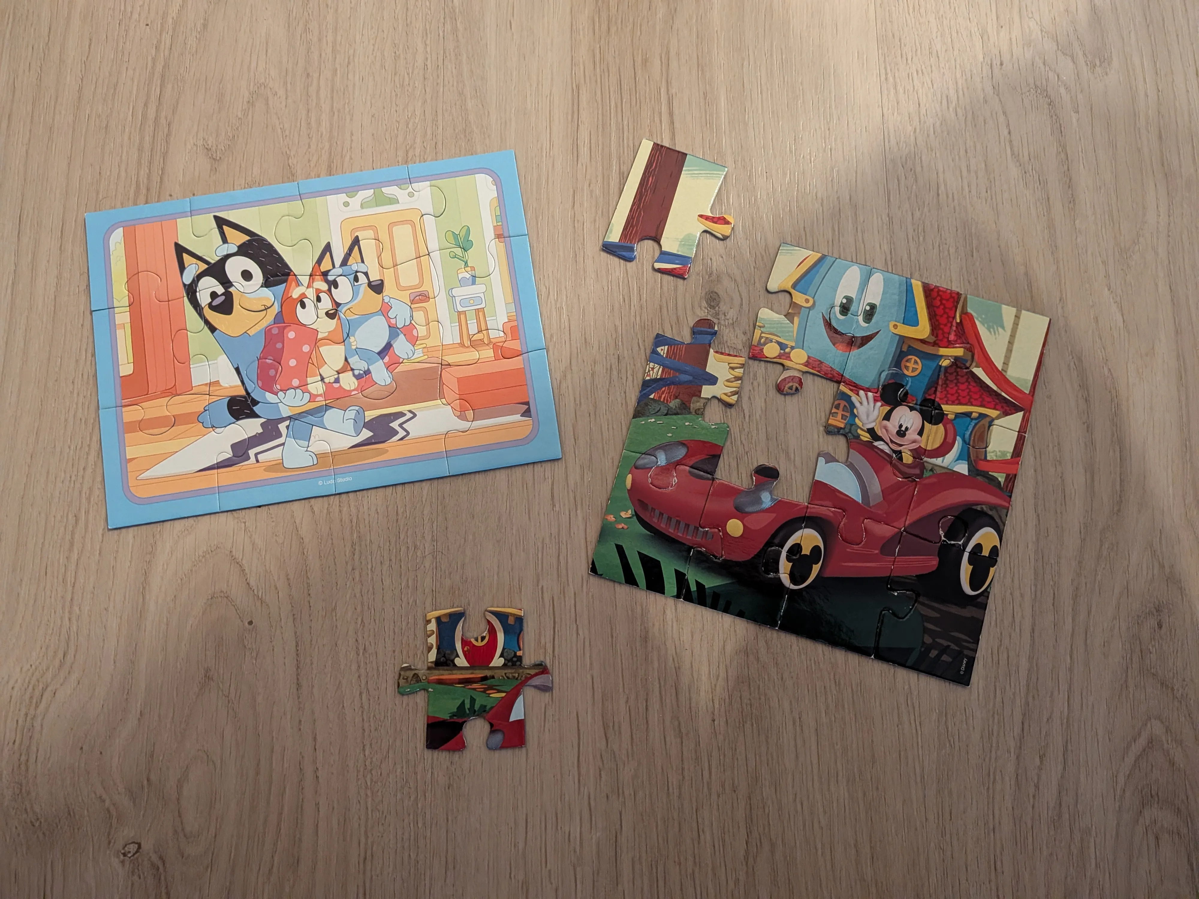 Dos puzzles infantiles casi terminados sobre el suelo, uno de Bluey y otro de Mickey, con tres piezas sueltas alrededor