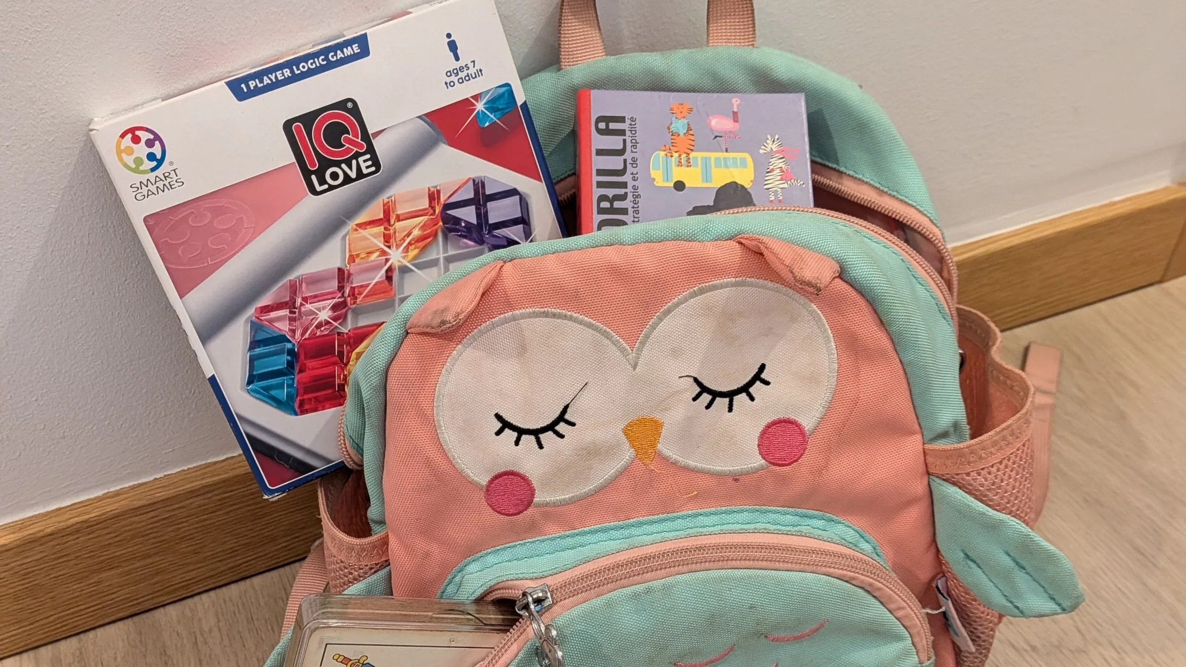 Una mochila infantil con forma de búho con el juego IQ Love, el Gorilla de Djeco y una baraja en su funda asomando por los bolsillos